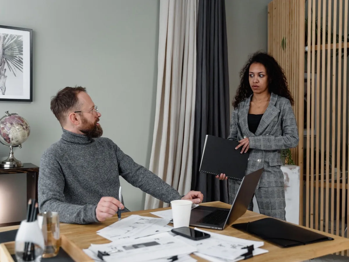 Three colleagues talk in an office, one standing with a laptop.