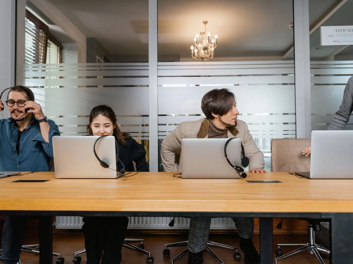 Several people work at laptops along a long table in an open office.