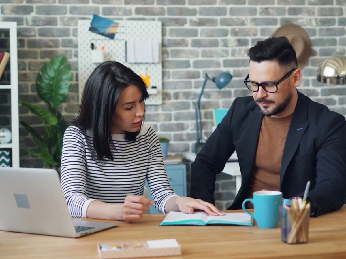 Two colleagues work at a laptop against a brick wall.